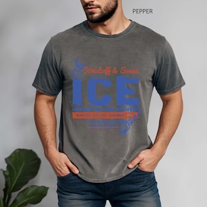 May include: A gray t-shirt with a blue and red graphic design. The design includes the word "ICE" in large blue letters, with additional text above and below. The shirt is a crew neck style.
