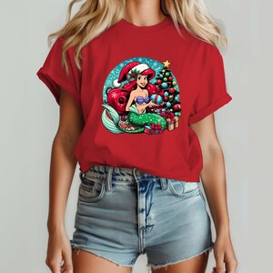 Puede incluir: Camiseta roja con un dise&ntilde;o de sirena de dibujos animados. La sirena tiene pelo rojo, una cola verde y lleva un gorro de Pap&aacute; Noel. Un &aacute;rbol de Navidad, regalos y un fondo nevado completan la escena festiva. El dise&ntilde;o es vibrante.