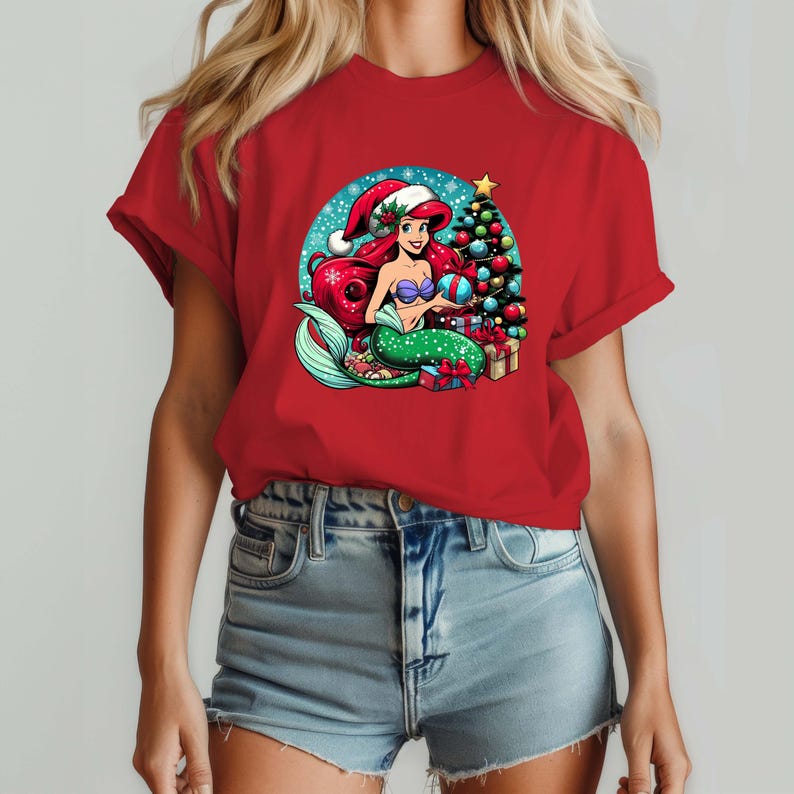 Puede incluir: Camiseta roja con un dise&ntilde;o de sirena de dibujos animados. La sirena tiene pelo rojo, una cola verde y lleva un gorro de Pap&aacute; Noel. Un &aacute;rbol de Navidad, regalos y un fondo nevado completan la escena festiva. El dise&ntilde;o es vibrante.