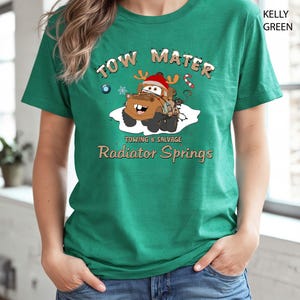 May include: Kelly green t-shirt featuring a cartoon image of Tow Mater from the movie Cars, wearing a Santa hat and reindeer antlers. The text "TOW MATER" and "Towing & Salvage, Radiator Springs" are printed above and below the image.
