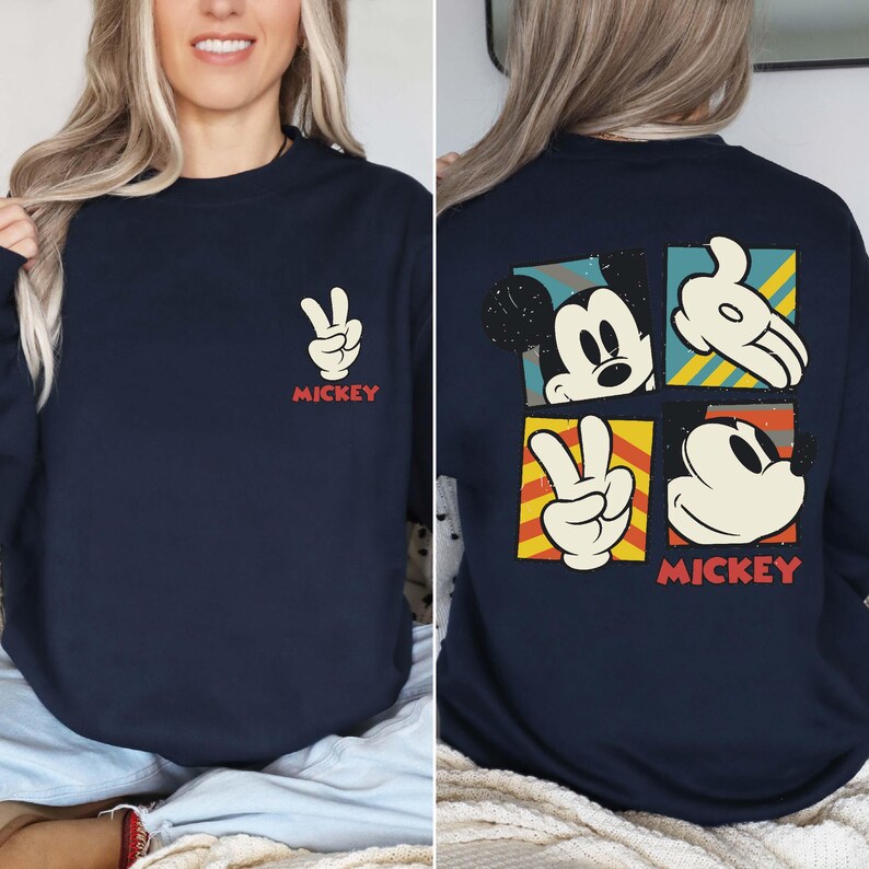 May include: Navy blue sweatshirt featuring Mickey Mouse-themed graphics. The front displays a peace sign hand gesture and the word "MICKEY" in red. The back showcases Mickey's face and hands in colorful squares, with "MICKEY" at the bottom.