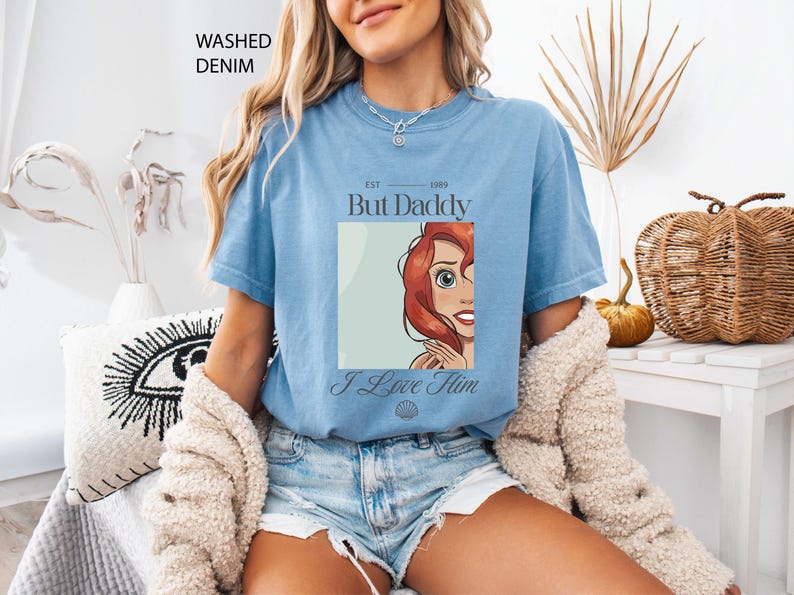 May include: Light blue t-shirt with a graphic of a cartoon character and the text "But Daddy I Love Him". The shirt is made of a soft material and is a washed denim color.
