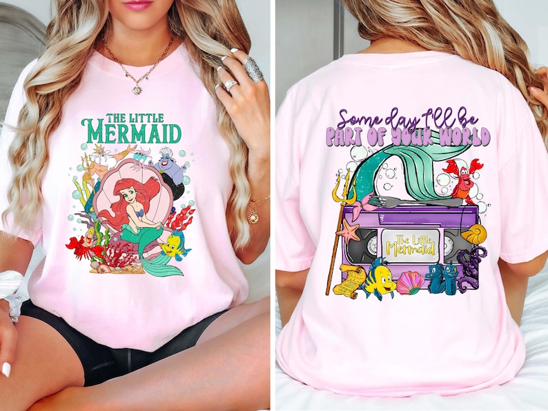 May include: Light pink t-shirt featuring a colorful graphic from "The Little Mermaid." The front design includes Ariel in a clam shell, with text "THE LITTLE MERMAID." The back design shows a VHS tape with the text "The Little Mermaid" and the phrase "Some day I'll be part of your world."