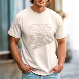 May include: A cream-colored short-sleeve t-shirt featuring a light brown graphic print of the Millennium Falcon spaceship centered on the chest, positioned below the Star Wars logo in a matching light brown outline style.