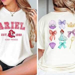 Może przedstawiać: Kremowa koszulka z napisem "ARIEL" w czerwonym stylu uniwersyteckim, z wizerunkiem Ariel i napisem "EST. 1989 DISNEY PRINCESS." Tył przedstawia ilustracje z motywem Ariel: kokardy, muszle, koronę i tr&oacute;jząb.