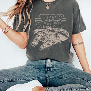 May include: A charcoal gray short-sleeve t-shirt featuring a beige line-art graphic of the Millennium Falcon spaceship and the Star Wars logo. The shirt is labeled as pepper color and is paired with blue denim jeans and white slip-on sneakers.