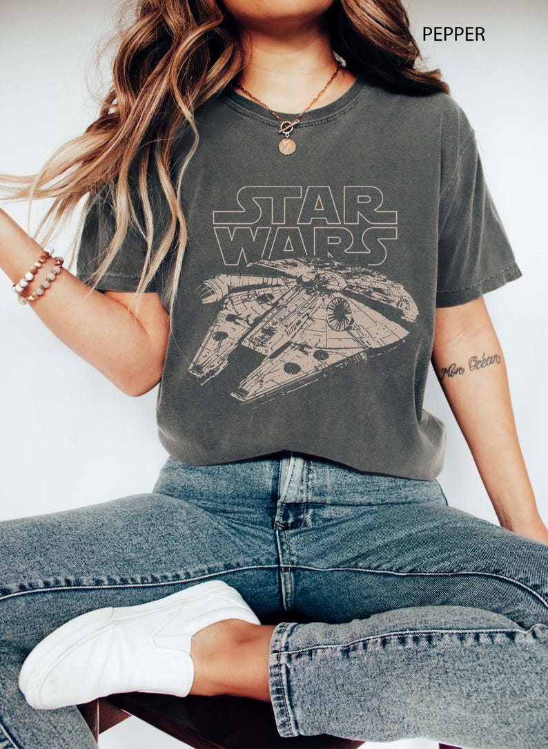 May include: A charcoal gray short-sleeve t-shirt featuring a beige line-art graphic of the Millennium Falcon spaceship and the Star Wars logo. The shirt is labeled as pepper color and is paired with blue denim jeans and white slip-on sneakers.