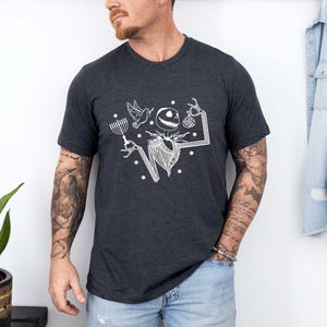 Op de afbeelding: Donkergrijs t-shirt met een witte afbeelding van een skeletachtig personage met een menora, duif en ornament. Het ontwerp bevat sterren en een gestileerd frame. Het shirt is gemaakt van zacht materiaal.