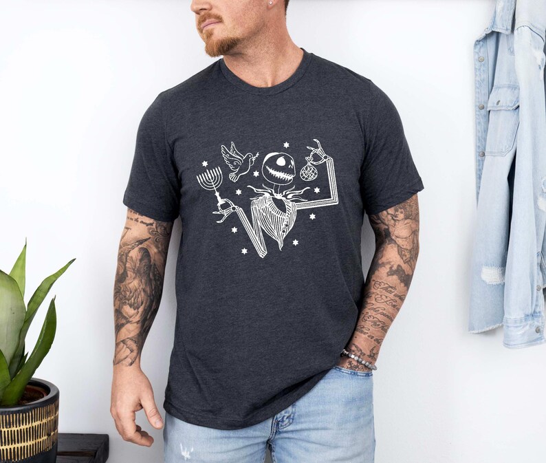 Op de afbeelding: Donkergrijs t-shirt met een witte afbeelding van een skeletachtig personage met een menora, duif en ornament. Het ontwerp bevat sterren en een gestileerd frame. Het shirt is gemaakt van zacht materiaal.