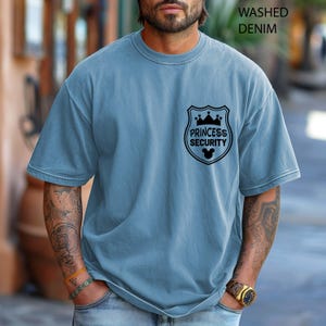 May include: A light blue, washed denim t-shirt with a black graphic on the chest that reads "Princess Security" inside a shield with a crown and Mickey Mouse ears. The shirt has short sleeves and a crew neck.