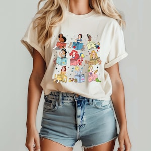 May include: Cream-colored t-shirt featuring a colorful graphic of various princesses reading books. Each princess is depicted with unique details, surrounded by small animals and stars. The design is printed on a soft, casual tee.