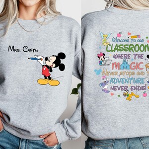 K&ouml;nnte beinhalten: Hellgraues Sweatshirt mit Mickey Mouse, der mit einem blauen Stift zeichnet. Der Text "Mrs. Cantu" steht &uuml;ber Mickey. Auf der R&uuml;ckseite des Sweatshirts steht "Welcome to our classroom where the magic never stops and the adventure never ends!"