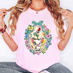 Puede incluir: Camiseta rosa claro con una ilustraci&oacute;n de dibujos animados de un cerdo y un gallo dentro de un marco ovalado floral. El dise&ntilde;o incluye una cinta turquesa y flores coloridas. La camiseta es de color rosa claro.