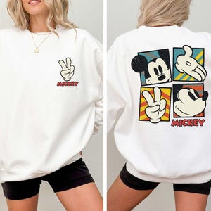 May include: White sweatshirt featuring a Mickey Mouse design. The front displays a peace sign hand gesture with the word "Mickey" in red. The back has four squares with Mickey Mouse images and the word "Mickey".