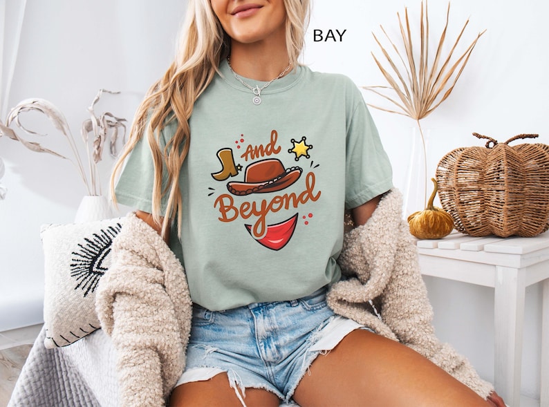 Może przedstawiać: Szałwiowa zielona koszulka z grafiką przedstawiającą kapelusz kowbojski, but i napisem "And Beyond". Projekt zawiera gwiazdę szeryfa i czerwony element. Koszulka jest połączona z dżinsowymi szortami.