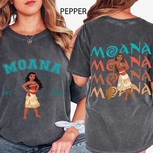 May include: Charcoal grey t-shirt with the word "MOANA" in teal, and an illustration of Moana. The back of the shirt repeats "MOANA" in teal, orange, and yellow, with a Moana illustration. The shirt is a crew neck.