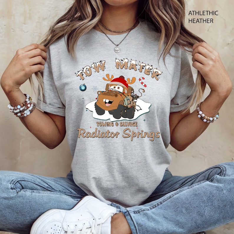 May include: Athletic heather gray t-shirt featuring a cartoon image of Tow Mater from the movie Cars, wearing a Santa hat and reindeer antlers. The text reads "Tow Mater Towing & Salvage Radiator Springs".