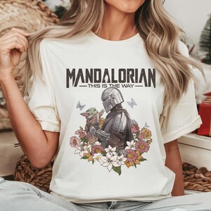 Puede incluir: Camiseta color crema con el texto "MANDALORIAN - THIS IS THE WAY". El gr&aacute;fico representa al Mandaloriano y a Grogu rodeados de flores y mariposas. El dise&ntilde;o est&aacute; impreso en una camiseta suave de manga corta.