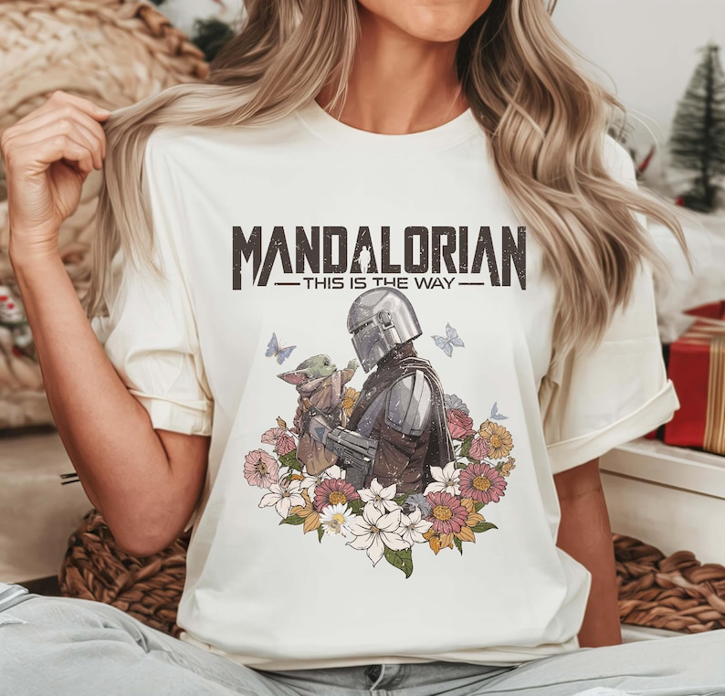 Puede incluir: Camiseta color crema con el texto "MANDALORIAN - THIS IS THE WAY". El gr&aacute;fico representa al Mandaloriano y a Grogu rodeados de flores y mariposas. El dise&ntilde;o est&aacute; impreso en una camiseta suave de manga corta.