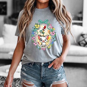 Puede incluir: Camiseta gris con un gr&aacute;fico colorido de un cerdo y un gallo de dibujos animados dentro de un marco ovalado floral. El dise&ntilde;o incluye una cinta azul claro y varias flores. La camiseta es de cuello redondo.