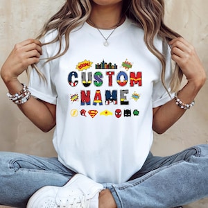 May include: White t-shirt featuring the words "CUSTOM NAME" in colorful, comic-book style lettering. Superhero symbols and sound effects like "BOOM!" and "ZOOM!" are also included in the design. The shirt is a casual, everyday wear item.