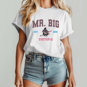 Puede incluir: Camiseta blanca con el texto "MR. BIG" en morado y un personaje de mapache de dibujos animados de Zootopia. La camiseta tambi&eacute;n presenta el texto "EST. 2016" y "ZOOTOPIA" en rosa. La modelo lleva pantalones cortos vaqueros azules.