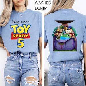 K&ouml;nnte beinhalten: Ein verwaschenes, denimblaues T-Shirt mit dem Disney Pixar Toy Story 5 Logo auf der Vorderseite in Gelb, Rot und Blau. Die R&uuml;ckseite des Shirts zeigt eine Grafik eines violetten Rucksacks, gef&uuml;llt mit verschiedenen animierten Spielzeugfiguren und einem gr&uuml;nen Frosch.