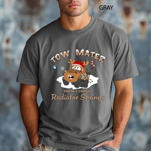 May include: Gray t-shirt featuring a cartoon image of Tow Mater from the movie Cars, wearing a Santa hat and reindeer antlers. The text "TOW MATER" is above the image, and "Towing & Salvage, Radiator Springs" is below.