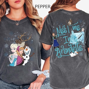 May include: Dark gray t-shirt featuring characters from the movie Frozen. The front of the shirt displays Elsa, Anna, Olaf, and Sven. The back of the shirt has the text "Meet me in Arendelle" with additional characters and snowflakes.