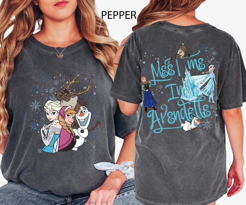 May include: Dark gray t-shirt featuring characters from the movie Frozen. The front of the shirt displays Elsa, Anna, Olaf, and Sven. The back of the shirt has the text "Meet me in Arendelle" with additional characters and snowflakes.