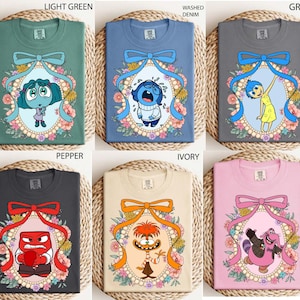 May include: T-shirts in light green, washed denim, grey, pepper, ivory, and pink. Each shirt features a cartoon character inside a floral frame with a bow. The characters include Joy, Sadness, Anger, and others.