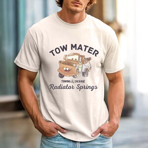 May include: Cream-colored t-shirt featuring a cartoon image of Tow Mater from the movie Cars. The graphic includes the text "TOW MATER" above the image, and "TOWING & SALVAGE" and "Radiator Springs" below. The shirt has short sleeves.
