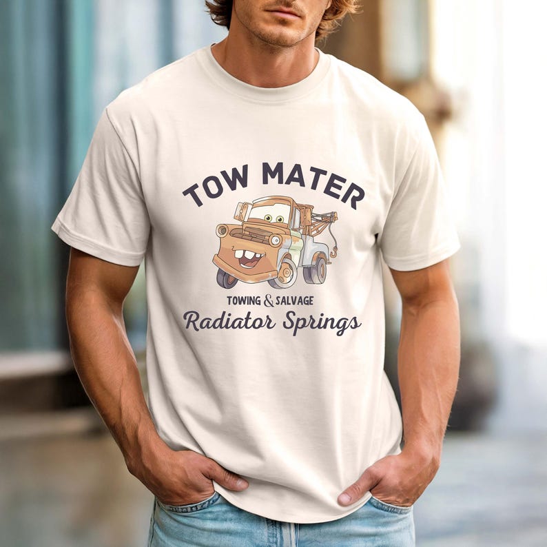 May include: Cream-colored t-shirt featuring a cartoon image of Tow Mater from the movie Cars. The graphic includes the text "TOW MATER" above the image, and "TOWING & SALVAGE" and "Radiator Springs" below. The shirt has short sleeves.