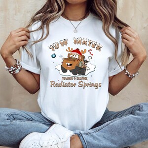 May include: White t-shirt featuring a cartoon image of Tow Mater from the movie Cars, wearing a Santa hat and antlers. The text "Tow Mater" and "Radiator Springs" are printed above and below the image, with additional text "Towing & Salvage".
