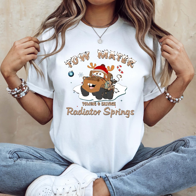 May include: White t-shirt featuring a cartoon image of Tow Mater from the movie Cars, wearing a Santa hat and antlers. The text "Tow Mater" and "Radiator Springs" are printed above and below the image, with additional text "Towing & Salvage".