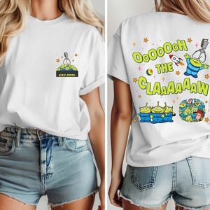 May include: White t-shirt featuring a cartoon design of the Little Green Men from Toy Story. The front has a small graphic with the text "EST.1995". The back displays the phrase "OooOoooh the Claaaaaaw" with additional graphics.