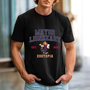 Puede incluir: Camiseta negra con la inscripci&oacute;n "MAYOR LIONHEART" y "ZOOTOPIA" en una fuente de estilo universitario. La camiseta presenta una ilustraci&oacute;n de un le&oacute;n sosteniendo un documento, con el texto "EST. 2016" debajo.