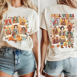 May include: Cream-colored t-shirt with a graphic featuring cartoon characters in autumn attire. The front design includes framed images of the characters, pumpkins, and fall leaves. The back of the shirt reads "FALL VIBES just a fall girl."