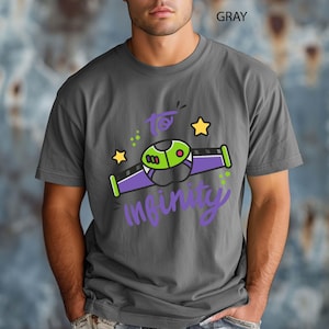 Może przedstawiać: Szara koszulka z grafiką przedstawiającą kresk&oacute;wkowy statek kosmiczny z napisem "to infinity" w fioletowym skrypcie. Statek kosmiczny ma zielone, fioletowe i ż&oacute;łte akcenty. U g&oacute;ry nadruk "GRAY".