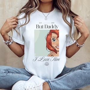 May include: White t-shirt with a graphic of a cartoon character with red hair and the text "But Daddy I Love Him" and "EST 1989". The character is in a light green rectangle. The shirt is a crew neck.