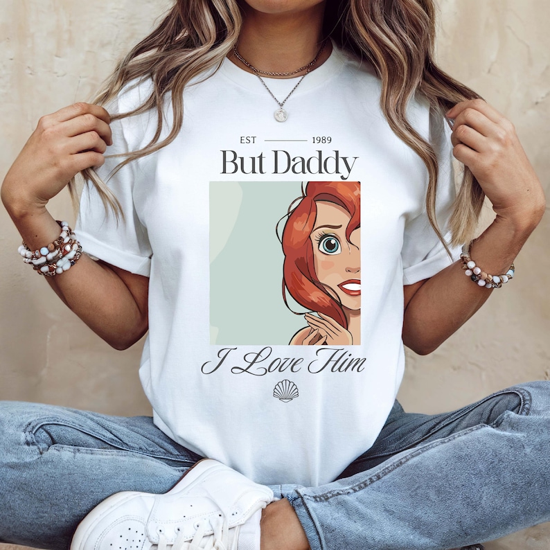 May include: White t-shirt with a graphic of a cartoon character with red hair and the text "But Daddy I Love Him" and "EST 1989". The character is in a light green rectangle. The shirt is a crew neck.