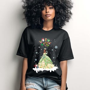 Op de afbeelding: Zwart t-shirt met een prinses in een groen en gele jurk die kerstballonnen vasthoudt. Een sneeuwman staat op een besneeuwde basis. Sneeuwvlokken zijn verspreid.