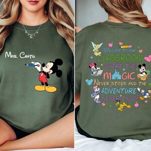 K&ouml;nnte beinhalten: Ein olivgr&uuml;nes Sweatshirt mit Mickey Mouse, der einen Bleistift h&auml;lt, und dem Namen "Mrs. Cantu" dar&uuml;ber. Auf der R&uuml;ckseite befindet sich ein farbenfrohes Design mit Disney-Figuren und dem Text "Welcome to our classroom where the magic never stops and the adventure never ends!"