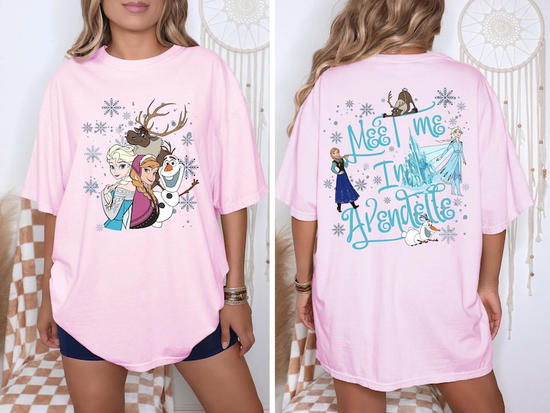 May include: Light pink t-shirt featuring characters from the animated film Frozen. The front displays Elsa, Anna, Olaf, and Sven with snowflakes. The back has the text "Meet me in Arendelle" with other characters and snowflakes.