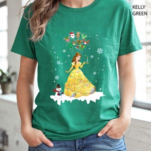 Op de afbeelding: Kelly Green t-shirt met een kerstthema ontwerp. De afbeelding toont een prinses in een gele jurk met een kaars, met kerstballonnen en een sneeuwpop. Het shirt is effen groen van kleur.