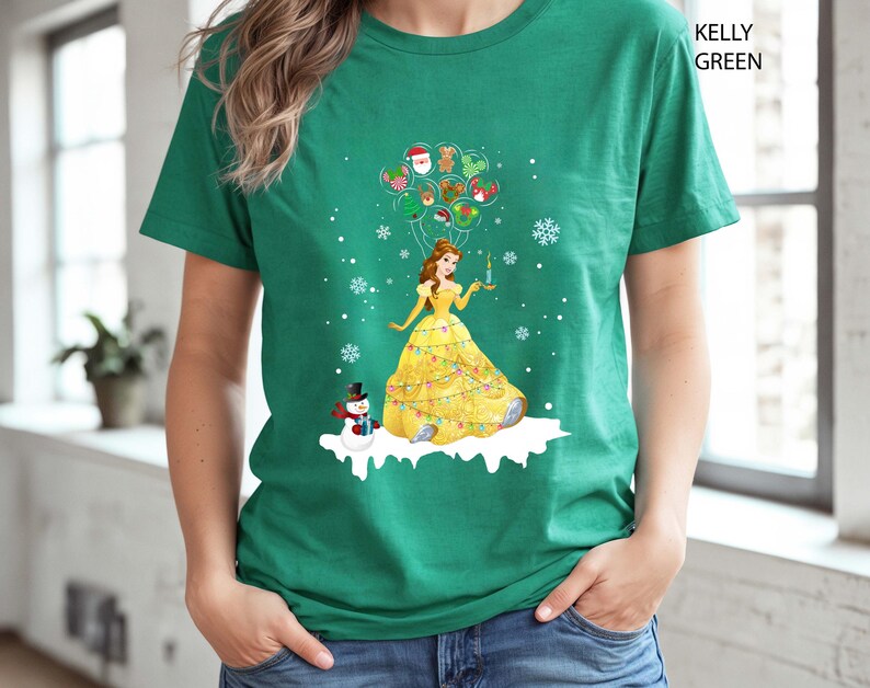 Op de afbeelding: Kelly Green t-shirt met een kerstthema ontwerp. De afbeelding toont een prinses in een gele jurk met een kaars, met kerstballonnen en een sneeuwpop. Het shirt is effen groen van kleur.