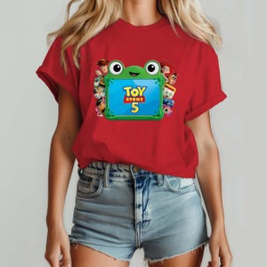 May include: A red short-sleeved t-shirt featuring a graphic of a green frog-shaped frame containing the text Toy Story 5. The frame is surrounded by various animated character figures including a cowboy, a cowgirl, and a potato-shaped character.