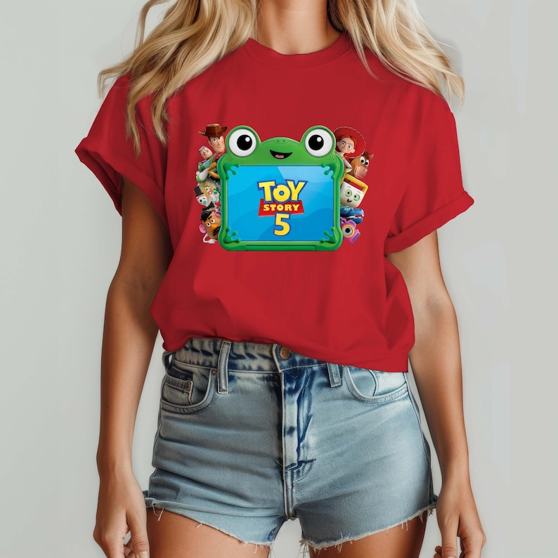 May include: A red short-sleeved t-shirt featuring a graphic of a green frog-shaped frame containing the text Toy Story 5. The frame is surrounded by various animated character figures including a cowboy, a cowgirl, and a potato-shaped character.