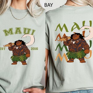 May include: A light green t-shirt with a cartoon illustration of Maui holding a fish hook. The front of the shirt has the word "MAUI" in green, with "EST 2016" below. The back of the shirt repeats "MAUI" in green, orange, and light green.