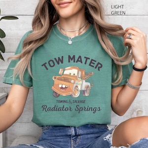May include: Light green t-shirt featuring a cartoon image of Tow Mater from the movie Cars. The text "TOW MATER" arches above the image, with "Towing & Salvage" and "Radiator Springs" below. The shirt is a comfortable, casual style.